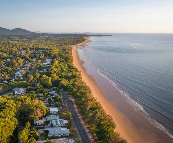 Aerial view of a serene coastline during sunset. The golden sandy beach curves alongside calm blue waters, bordered by lush green trees and a small coastal town.