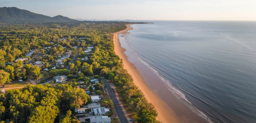Aerial view of a serene coastline during sunset. The golden sandy beach curves alongside calm blue waters, bordered by lush green trees and a small coastal town.