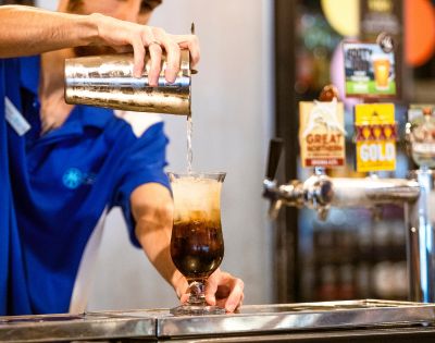 A bartender in a blue shirt pours a fizzy drink from a shaker into a tall glass filled with ice at a bar, evoking a lively, refreshing vibe.