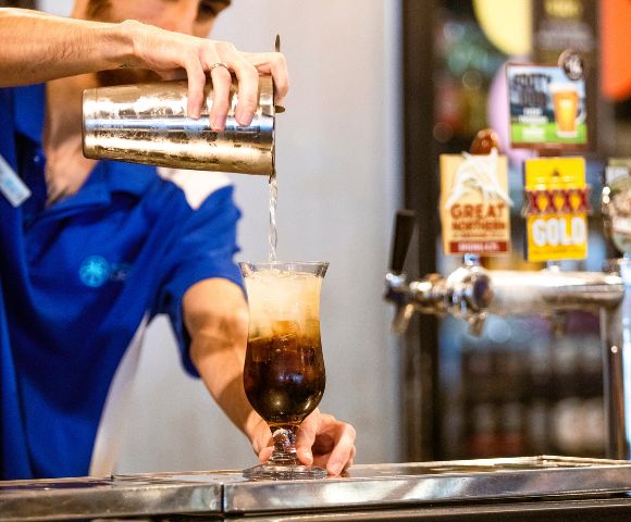 A bartender in a blue shirt pours a fizzy drink from a shaker into a tall glass filled with ice at a bar, evoking a lively, refreshing vibe.
