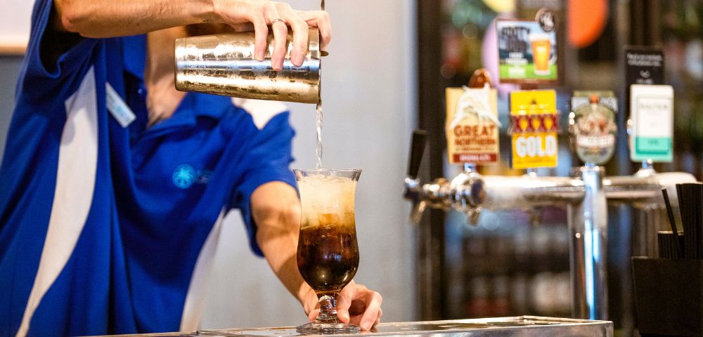 A bartender in a blue shirt pours a fizzy drink from a shaker into a tall glass filled with ice at a bar, evoking a lively, refreshing vibe.