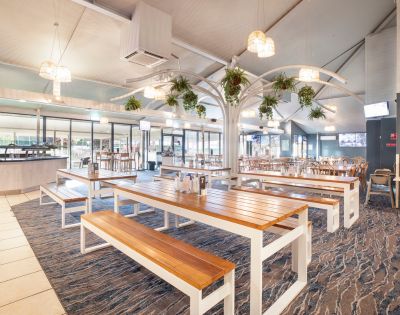 Spacious restaurant interior with wooden tables, benches, and hanging plants. Natural light and a modern, airy ambiance create a welcoming atmosphere.