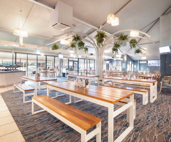 Spacious restaurant interior with wooden tables, benches, and hanging plants. Natural light and a modern, airy ambiance create a welcoming atmosphere.