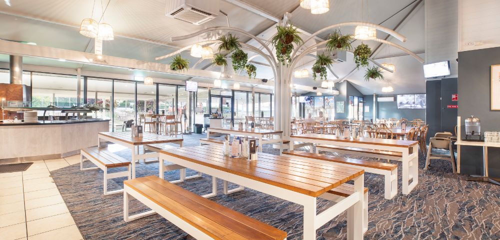 Spacious restaurant interior with wooden tables, benches, and hanging plants. Natural light and a modern, airy ambiance create a welcoming atmosphere.