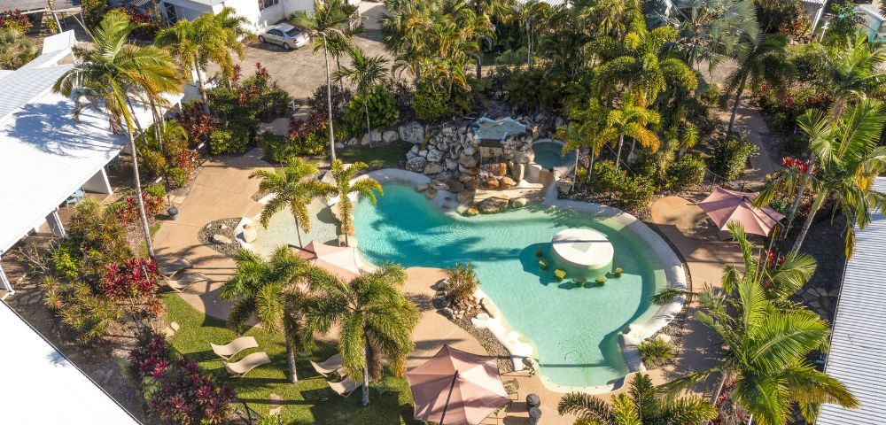 Aerial view of a tropical resort pool surrounded by lush palm trees and greenery. Lounge chairs and umbrellas create a relaxing, inviting atmosphere.