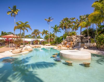 Tropical pool scene with clear blue water, surrounded by palm trees and rocks under a bright sky. Relaxing, inviting atmosphere.