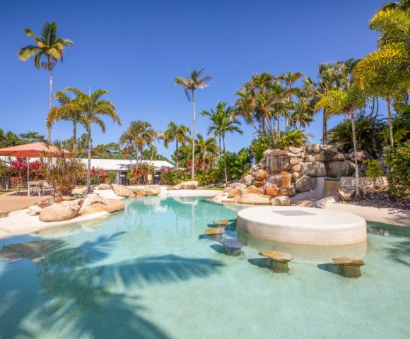 Tropical pool scene with clear blue water, surrounded by palm trees and rocks under a bright sky. Relaxing, inviting atmosphere.