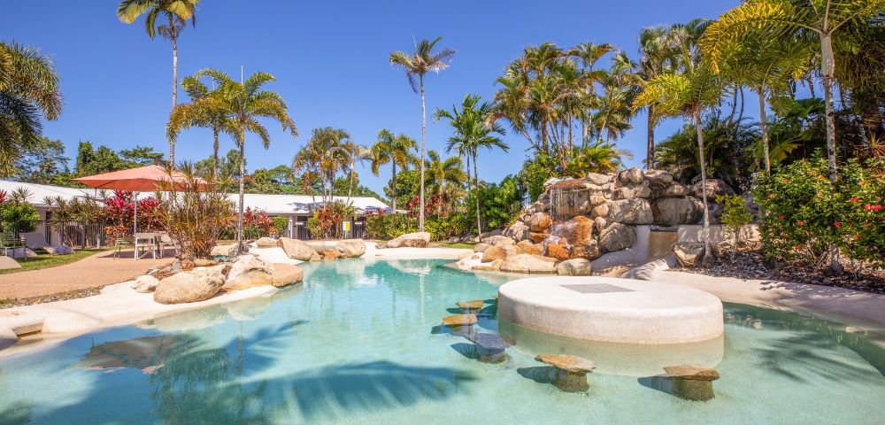 Tropical pool scene with clear blue water, surrounded by palm trees and rocks under a bright sky. Relaxing, inviting atmosphere.