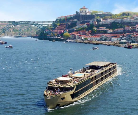 A large river cruise ship sails on a sunny day, passing under an arched bridge with a scenic hillside town in the background. The scene is tranquil and picturesque.