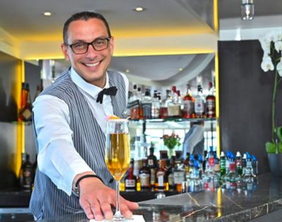 Smiling bartender in a striped vest presents a tall cocktail on a napkin at a modern bar. Liquor bottles and orchids enhance the elegant setting.
