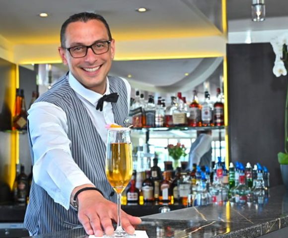 Smiling bartender in a striped vest presents a tall cocktail on a napkin at a modern bar. Liquor bottles and orchids enhance the elegant setting.