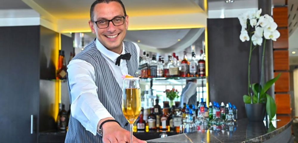 Smiling bartender in a striped vest presents a tall cocktail on a napkin at a modern bar. Liquor bottles and orchids enhance the elegant setting.
