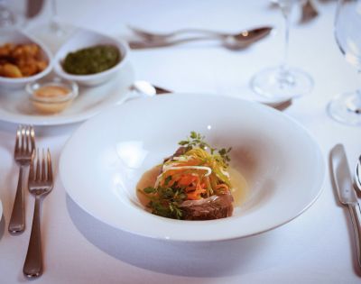 A gourmet dish with garnished meat and colorful vegetables is elegantly presented on a white plate. The table is set with silverware, wine glasses, and side dishes.