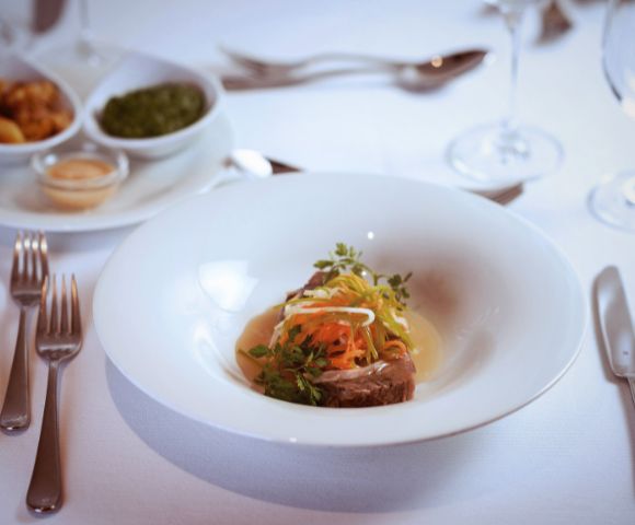 A gourmet dish with garnished meat and colorful vegetables is elegantly presented on a white plate. The table is set with silverware, wine glasses, and side dishes.