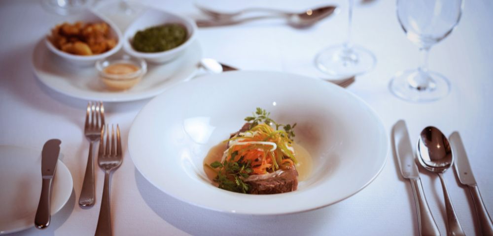 A gourmet dish with garnished meat and colorful vegetables is elegantly presented on a white plate. The table is set with silverware, wine glasses, and side dishes.
