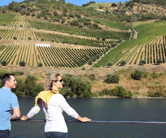 A couple enjoys a scenic view from a railing, overlooking a lush, sunlit vineyard on a hillside. The atmosphere is peaceful and picturesque.