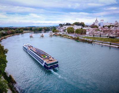 A large river cruise ship sails along a wide river past a town with historic buildings and a bridge. The sky is cloudy, and trees line the riverbank.