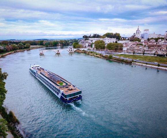A large river cruise ship sails along a wide river past a town with historic buildings and a bridge. The sky is cloudy, and trees line the riverbank.
