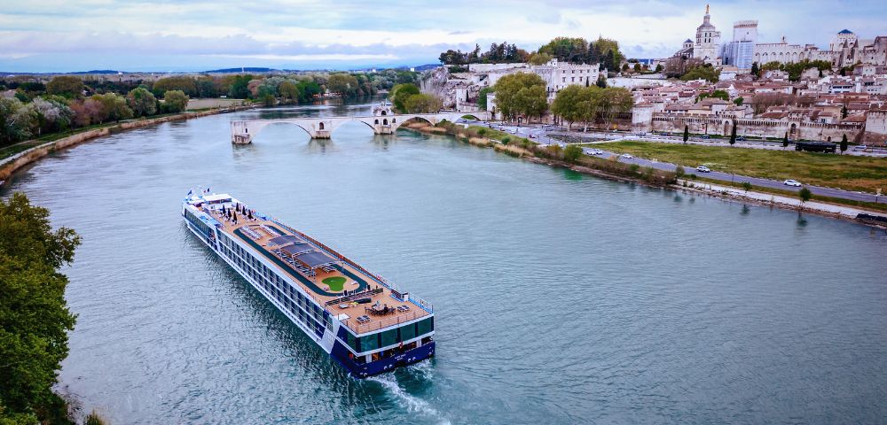A large river cruise ship sails along a wide river past a town with historic buildings and a bridge. The sky is cloudy, and trees line the riverbank.