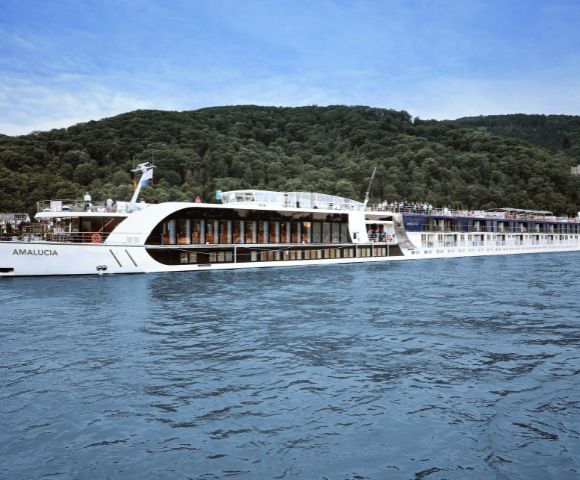 A large white river cruise ship sails on a calm body of water. Lush green hills are in the background under a bright blue sky. The scene is peaceful and serene.