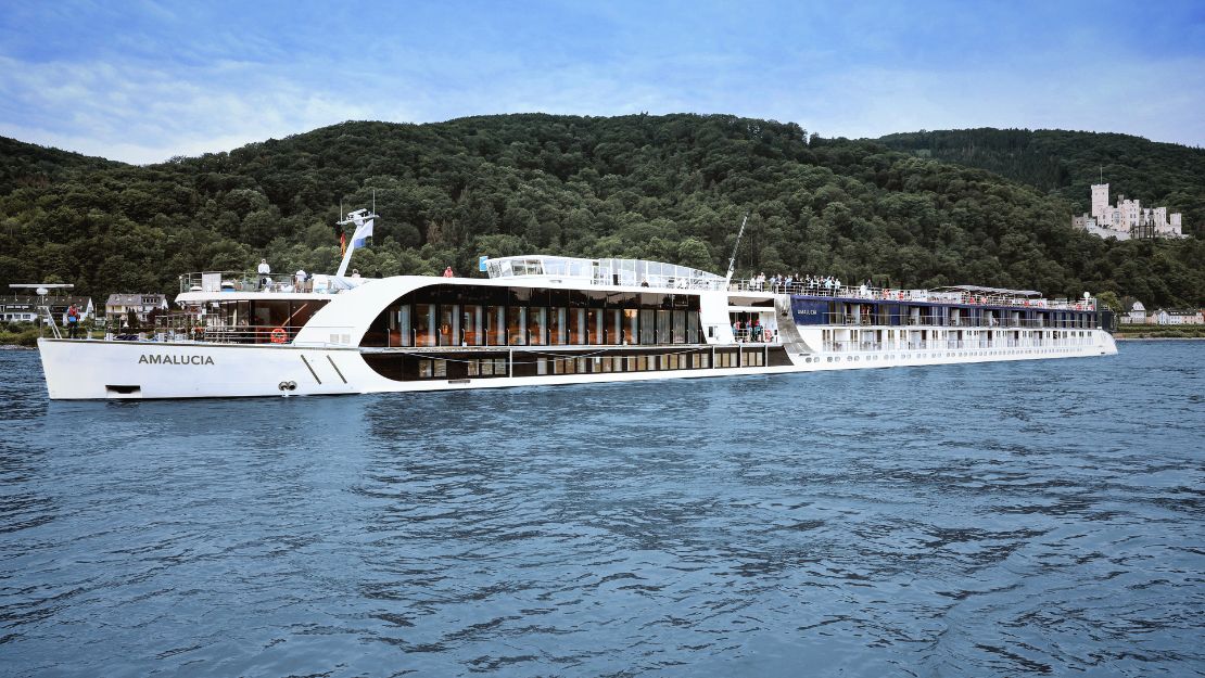 A large white river cruise ship sails on a calm body of water. Lush green hills are in the background under a bright blue sky. The scene is peaceful and serene.