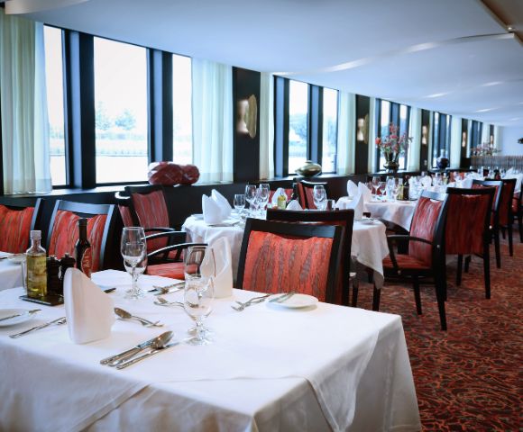 Luxurious restaurant with elegant decor, white tablecloths, red chairs, and soft lighting. Large windows provide natural light and a serene atmosphere.