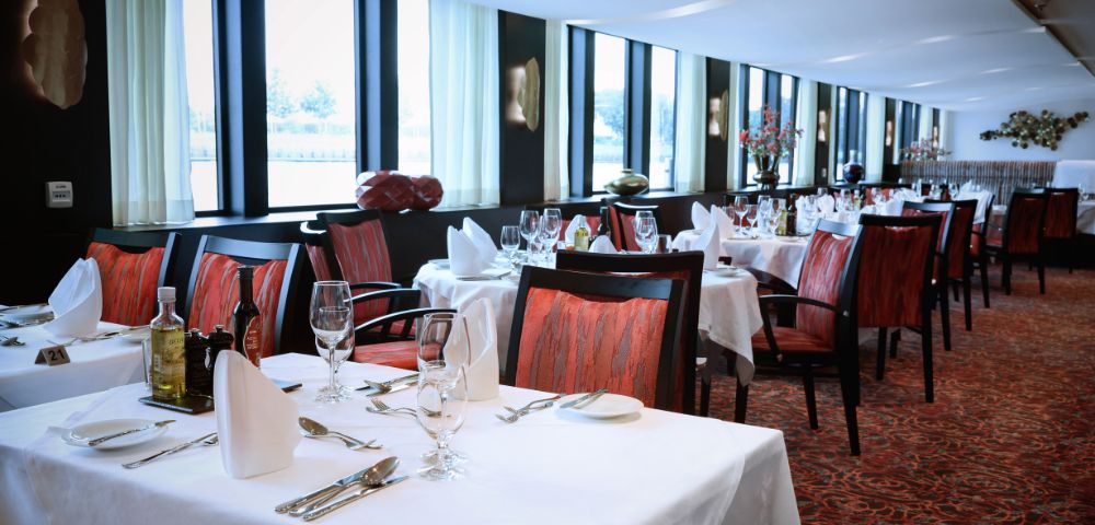 Luxurious restaurant with elegant decor, white tablecloths, red chairs, and soft lighting. Large windows provide natural light and a serene atmosphere.