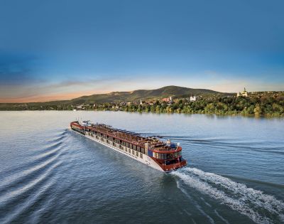 A large river cruise ship sails through calm blue water at sunset, creating gentle waves. Green hills and a distant town form the serene backdrop.