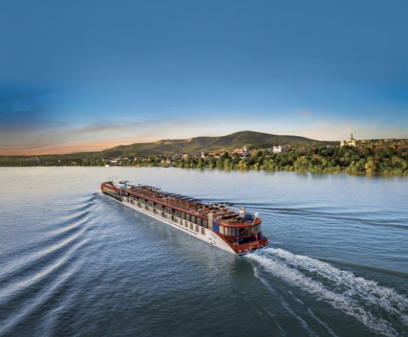 A large river cruise ship sails through calm blue water at sunset, creating gentle waves. Green hills and a distant town form the serene backdrop.