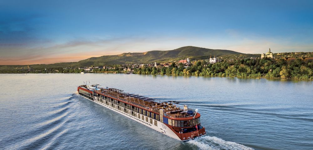 A large river cruise ship sails through calm blue water at sunset, creating gentle waves. Green hills and a distant town form the serene backdrop.