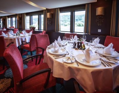 Elegant restaurant with large windows and scenic views. Round tables are set with white tablecloths, folded napkins, and sparkling glassware. Red chairs enhance the cozy ambiance.