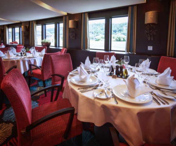 Elegant restaurant with large windows and scenic views. Round tables are set with white tablecloths, folded napkins, and sparkling glassware. Red chairs enhance the cozy ambiance.