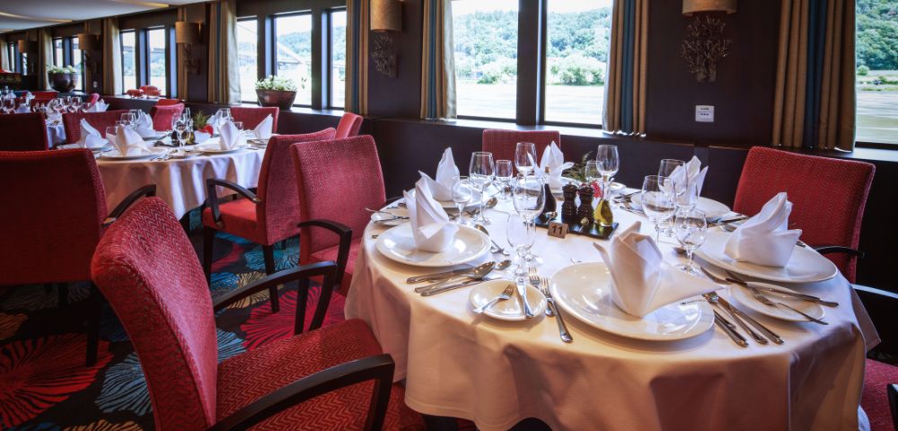 Elegant restaurant with large windows and scenic views. Round tables are set with white tablecloths, folded napkins, and sparkling glassware. Red chairs enhance the cozy ambiance.