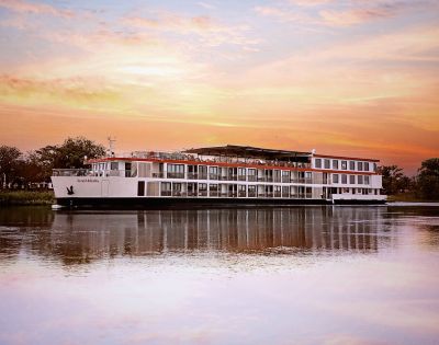 A large river cruise ship floats on calm water during a vibrant sunset, with hues of orange and pink in the sky. The scene is peaceful and serene.