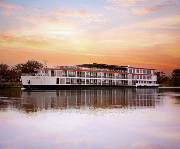 A large river cruise ship floats on calm water during a vibrant sunset, with hues of orange and pink in the sky. The scene is peaceful and serene.