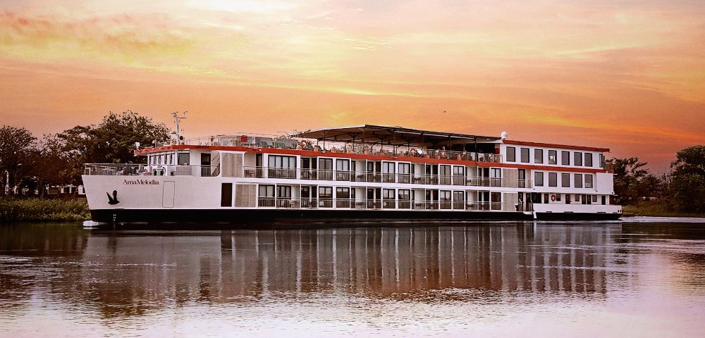 A large river cruise ship floats on calm water during a vibrant sunset, with hues of orange and pink in the sky. The scene is peaceful and serene.