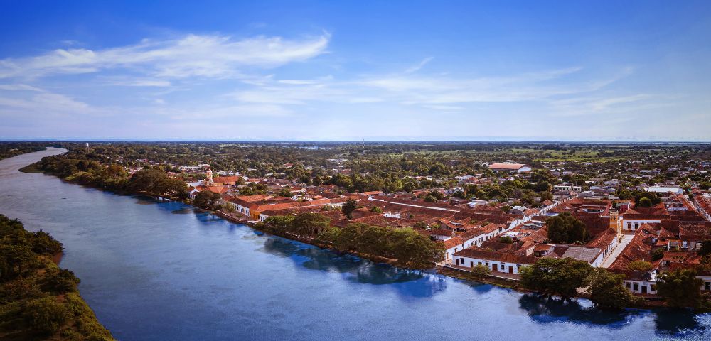 Aerial view of a historic town by a river, featuring red-tiled roofs and white colonial buildings. The scene conveys a serene and timeless atmosphere.