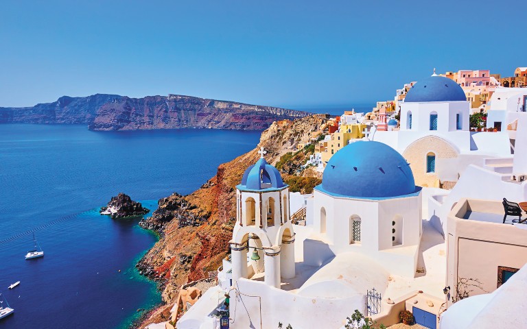 Scenic view of Santorini with white buildings and blue domes overlooking the Aegean Sea, with cliffs and a clear blue sky in the background.