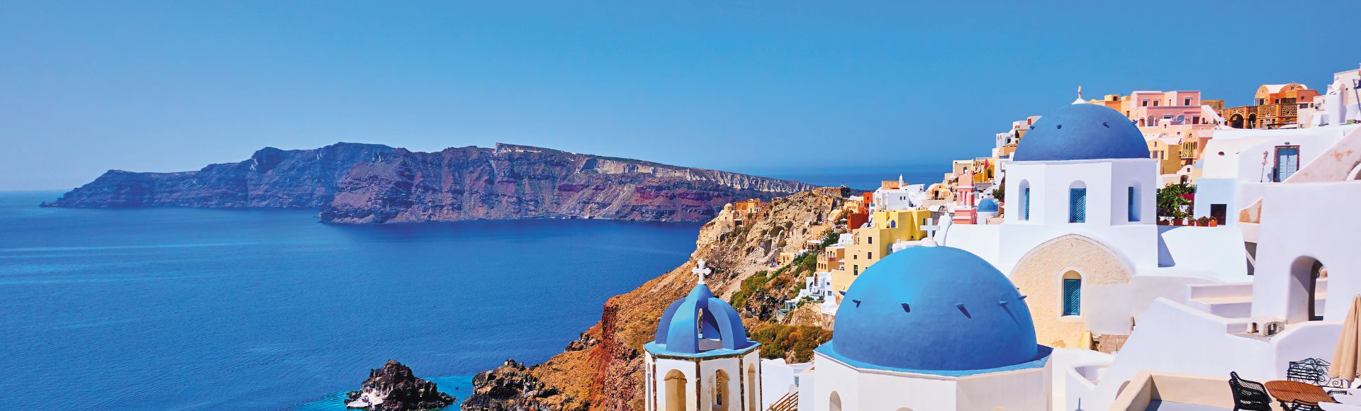 Scenic view of Santorini with white buildings and blue domes overlooking the Aegean Sea, with cliffs and a clear blue sky in the background.