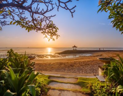 Sunrise over a tranquil beach, framed by leafy branches and lush plants, with a serene hut on the horizon, evoking calmness and peace.