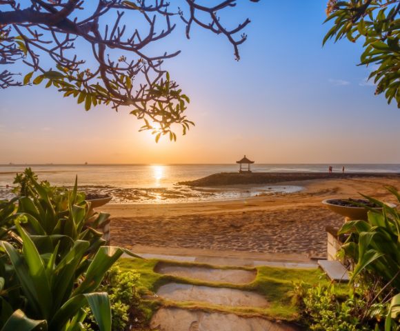 Sunrise over a tranquil beach, framed by leafy branches and lush plants, with a serene hut on the horizon, evoking calmness and peace.