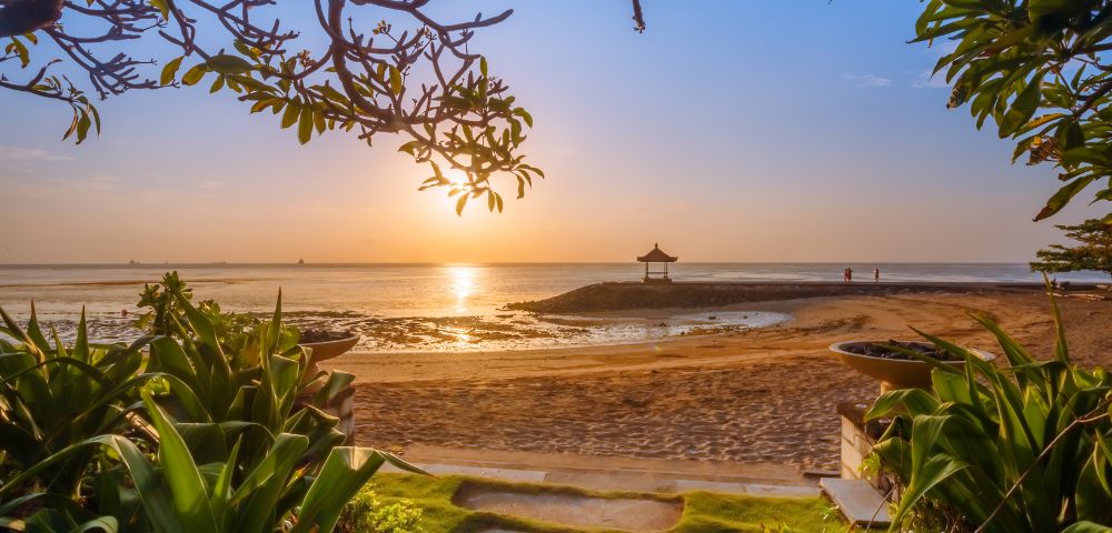 Sunrise over a tranquil beach, framed by leafy branches and lush plants, with a serene hut on the horizon, evoking calmness and peace.
