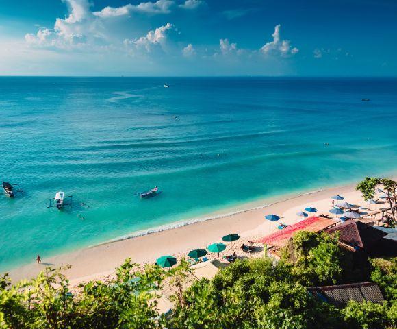 Scenic view of a tropical beach with turquoise water and gentle waves. Umbrellas and sun loungers line the sandy shore under a blue sky with clouds.