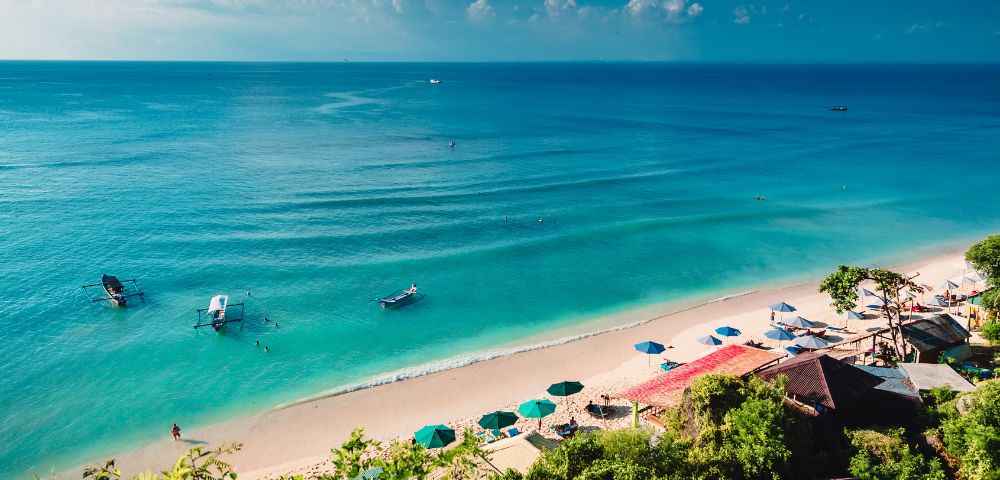 Scenic view of a tropical beach with turquoise water and gentle waves. Umbrellas and sun loungers line the sandy shore under a blue sky with clouds.