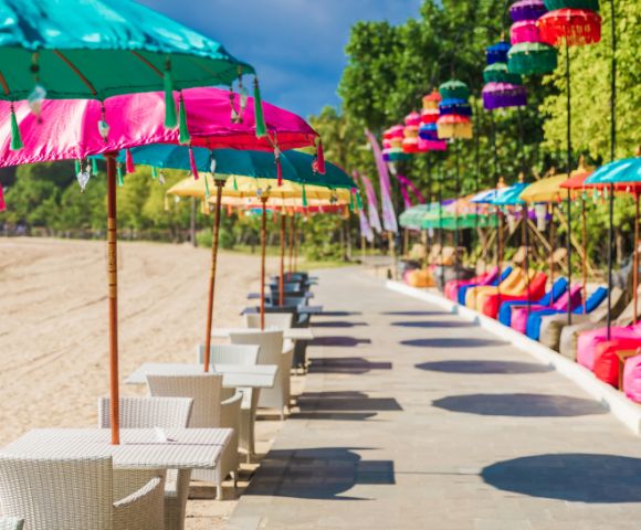 A vibrant beachside scene with colorful umbrellas and tables along a sunny promenade. Bright bean bags and lush trees line the background, evoking a tropical vibe.