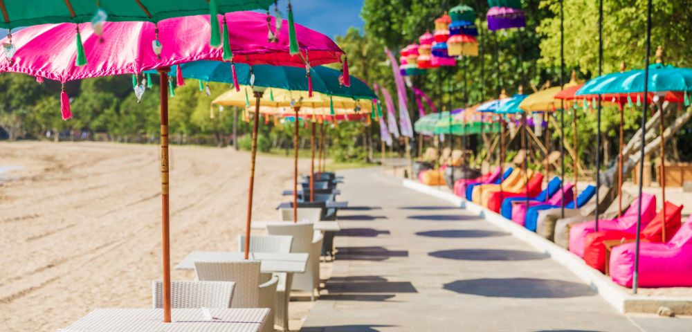A vibrant beachside scene with colorful umbrellas and tables along a sunny promenade. Bright bean bags and lush trees line the background, evoking a tropical vibe.