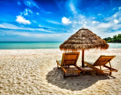 Sunny beach scene with two wooden lounge chairs under a straw umbrella on white sandy shore, overlooking serene turquoise ocean and vibrant blue sky.