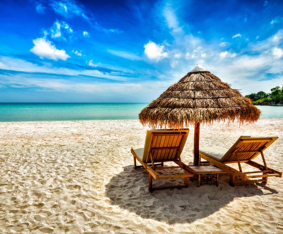 Sunny beach scene with two wooden lounge chairs under a straw umbrella on white sandy shore, overlooking serene turquoise ocean and vibrant blue sky.