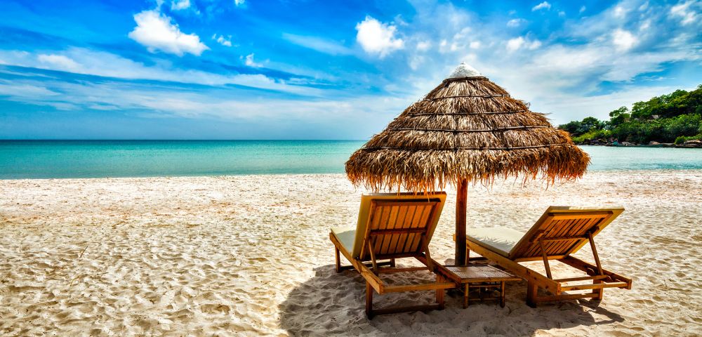 Sunny beach scene with two wooden lounge chairs under a straw umbrella on white sandy shore, overlooking serene turquoise ocean and vibrant blue sky.