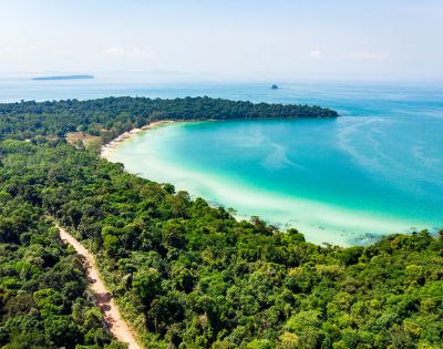 Aerial view of a lush green tropical forest bordering a turquoise bay with a sandy beach. The sky is clear, conveying a serene, idyllic atmosphere.